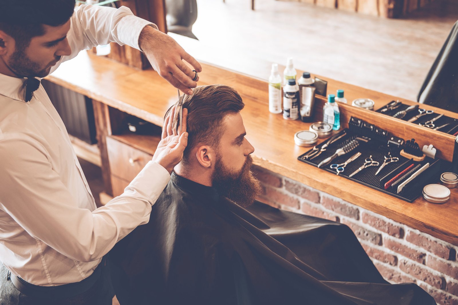 Haircut must be perfect. Top view of young bearded man getting haircut by hairdresser while sitting in chair at barbershop